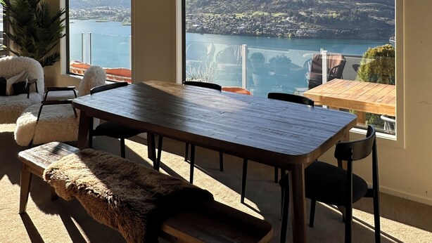 Un espace repas avec une grande table et des bancs, de grandes fenêtres offrant une vue sur les montagnes et des luminaires suspendus.