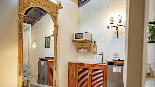 A room with a large ornate mirror, a wooden cabinet, a small refrigerator, and a bed with a patterned bedspread.