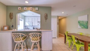 A kitchen with white cabinets, a microwave, and a dining area with a wooden table and bright green chairs. - Coconut Cove: ACROSS from beach + boardwalk + parking w/ Full Linen Service! (Ocean City)
