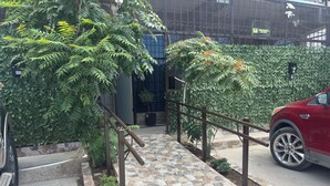 Uma trilha pavimentada leva à entrada de um prédio, rodeada por vegetação e um veículo vermelho estacionado perto.