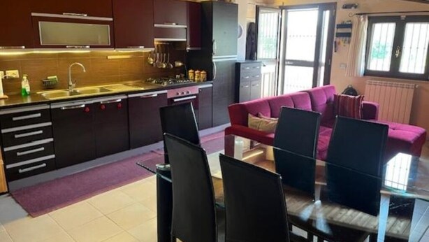 A modern kitchen with dark cabinetry, a gas stove, and a built-in oven. There is a dining area with a glass table and black chairs. A red sofa is visible in the background. - MODERN APARTMENT SURROUNDED BY GREENERY BUT A STONE'S THROW FROM THE SEA AND MOUNTAINS (Lazio)
