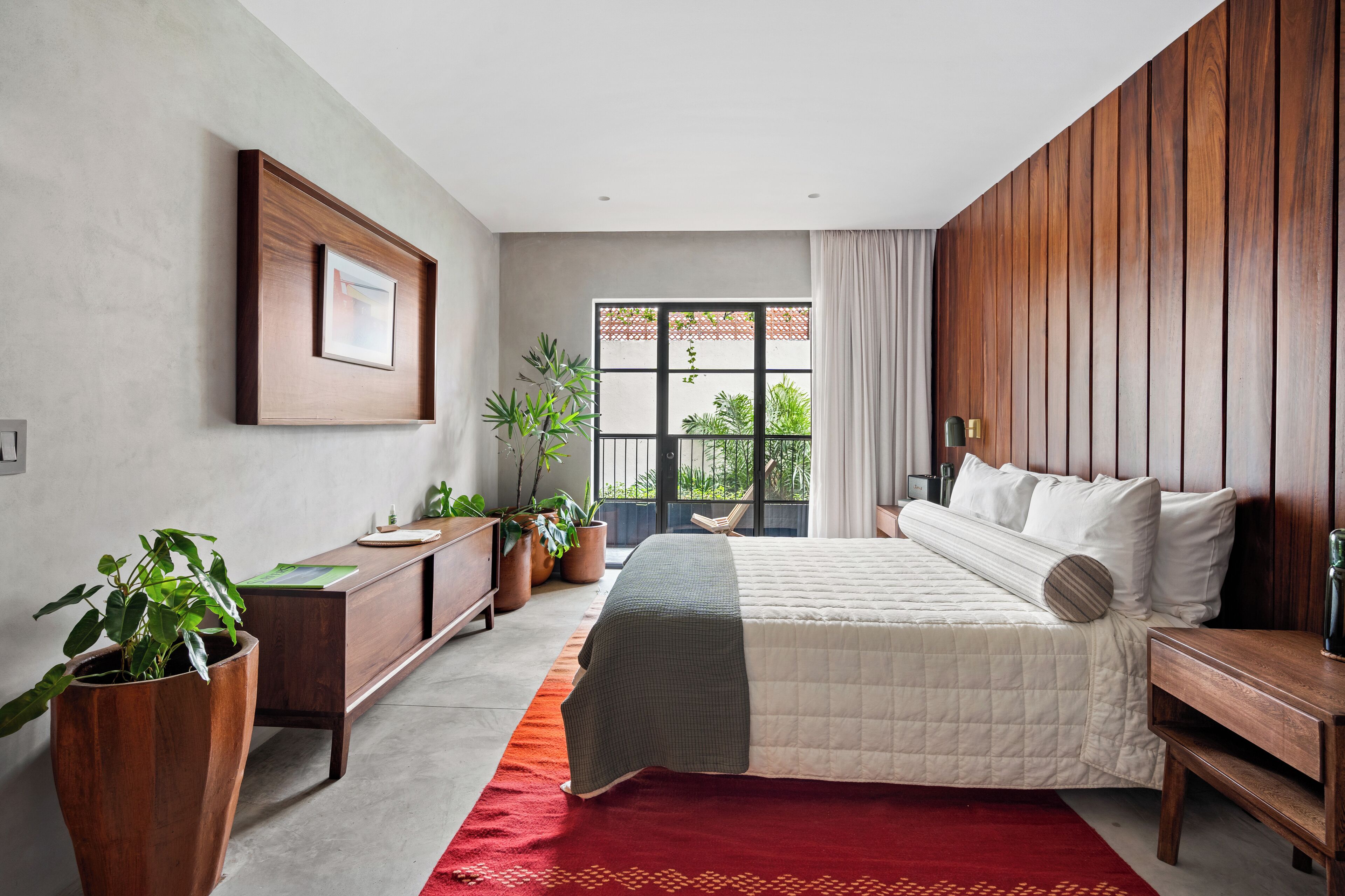 A bedroom with a bed, a wooden dresser, a desk, and potted plants.