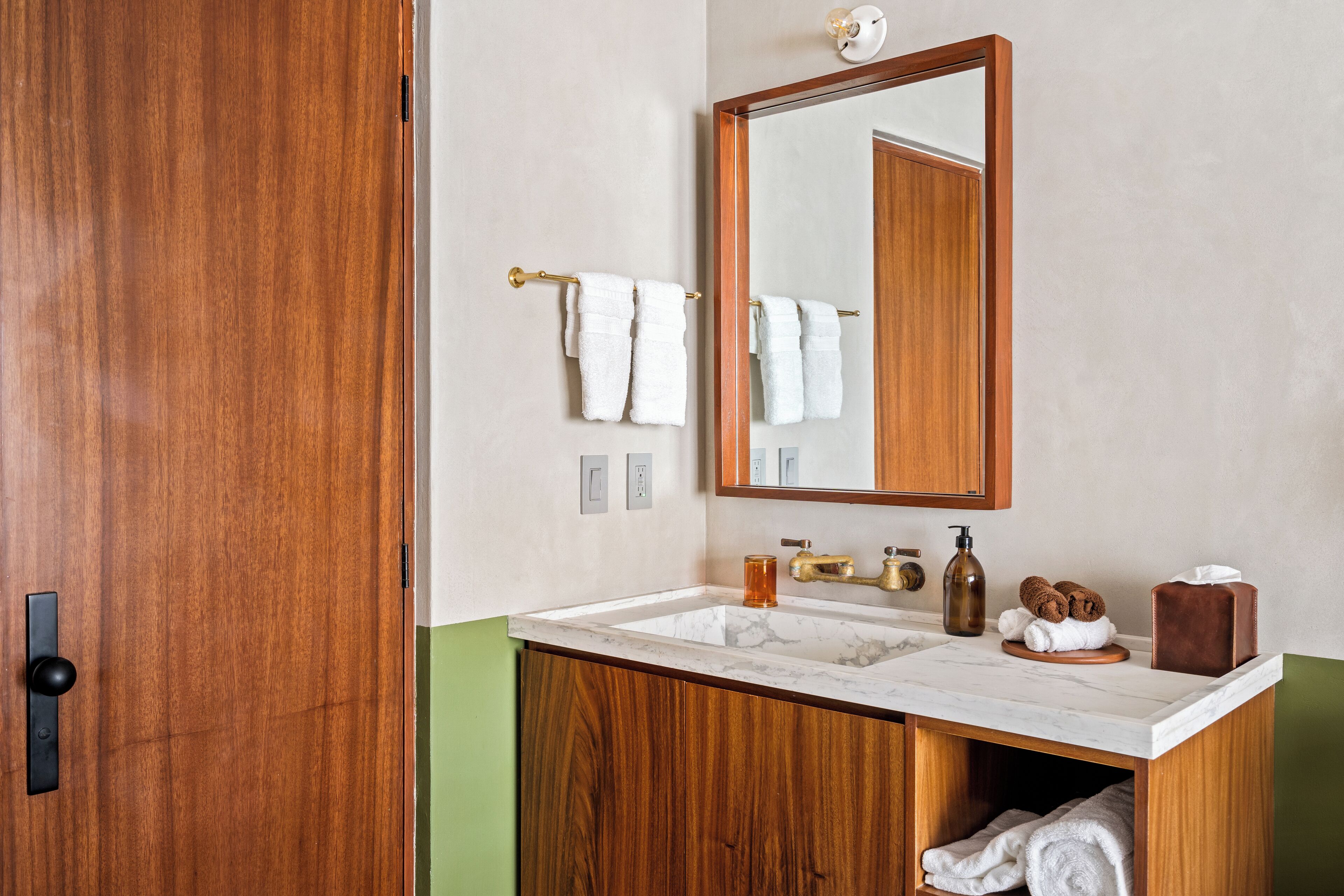Un baño con puerta de madera, lavamanos de mármol, espejo y perchero para toallas.