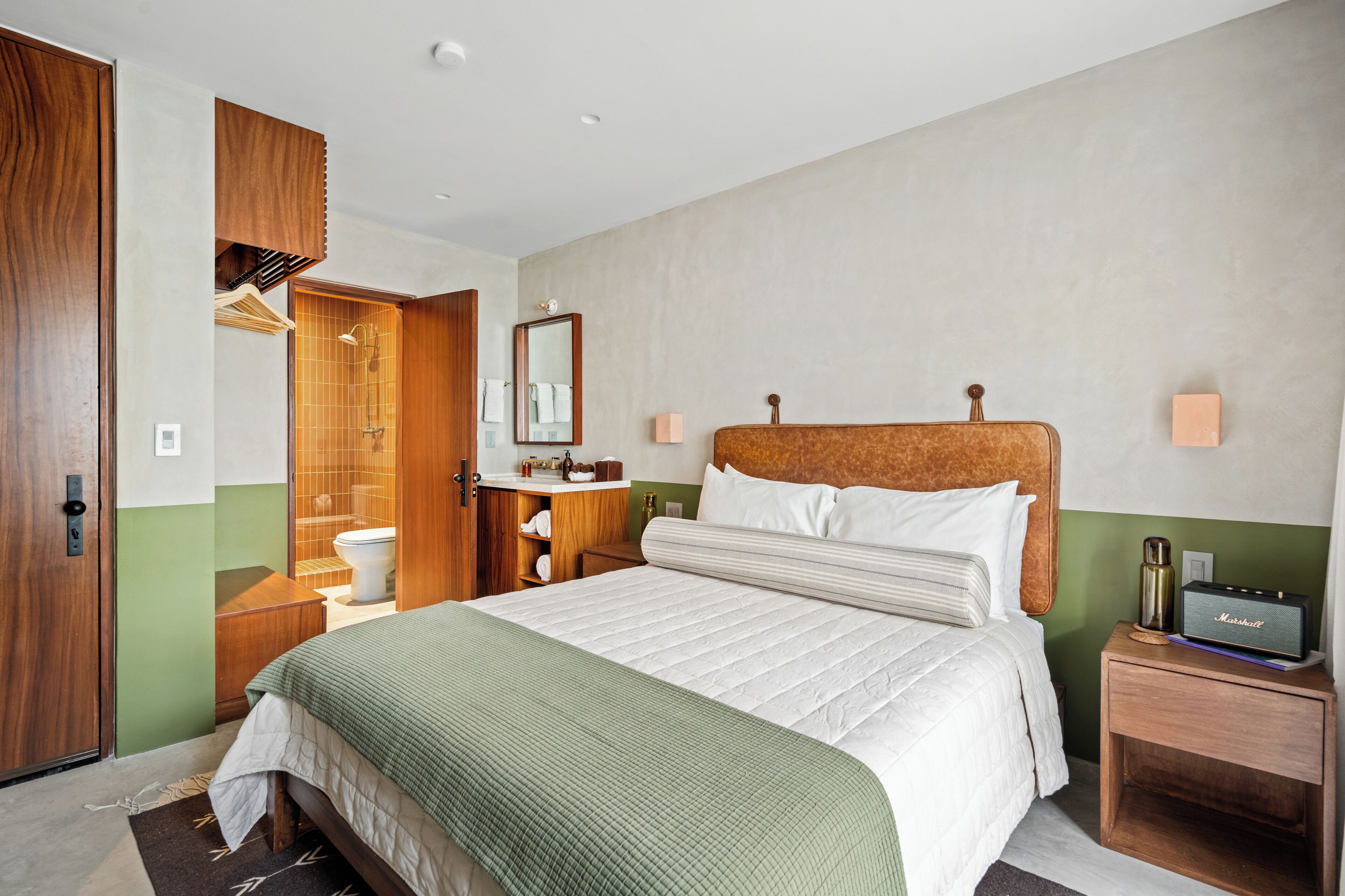 A hotel room with a large bed, wooden headboard, a bathroom with a shower and toilet, a small bedside table with a radio, and a green accent wall.