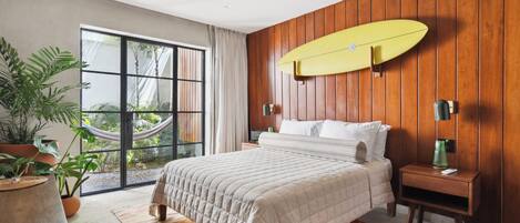 Een moderne slaapkamer met een groot bed, houten lambrisering, een surfplank en uitzicht op een tuin door een schuifdeur.
