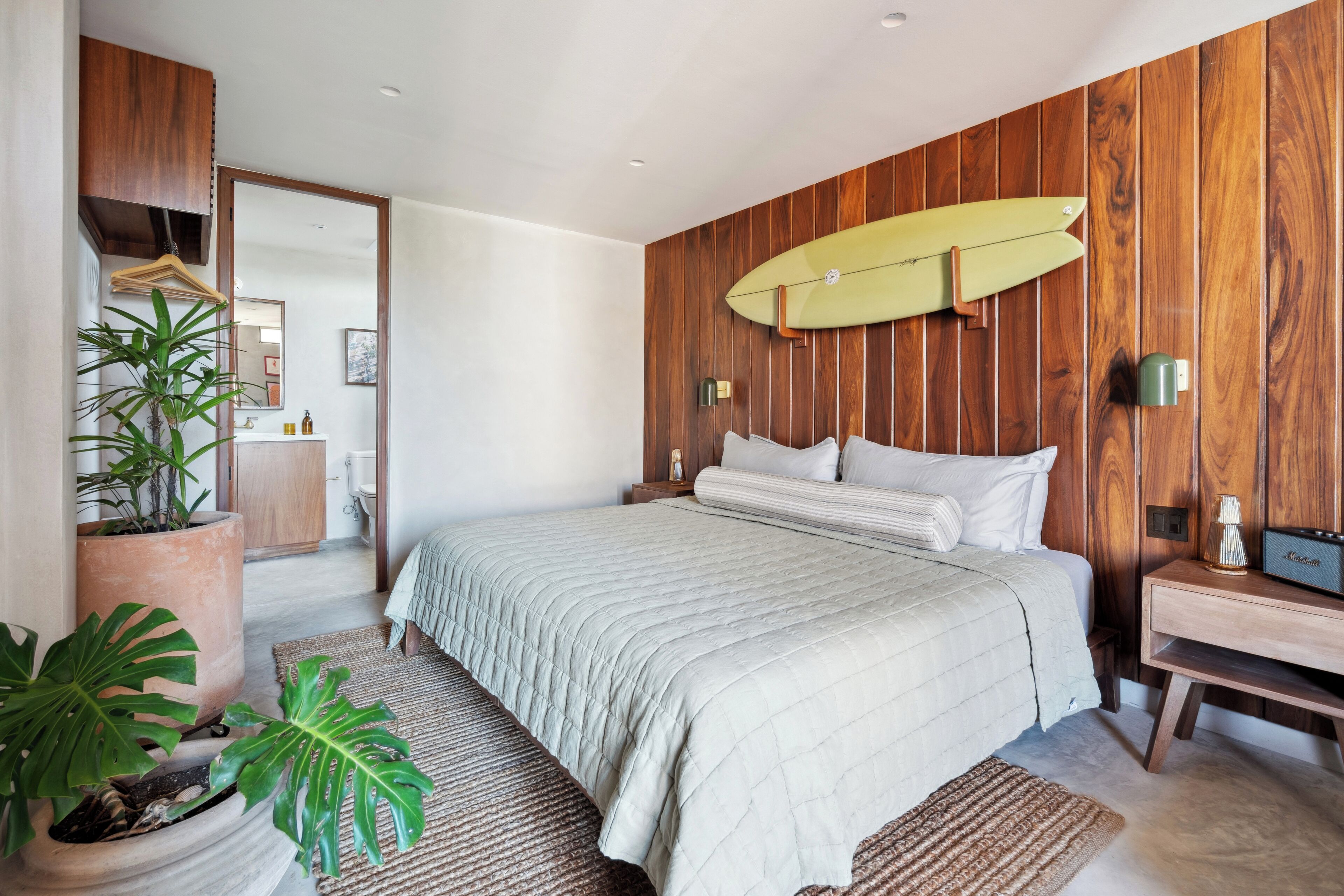 A bedroom with a large bed, wooden paneling, a surfboard, and potted plants.