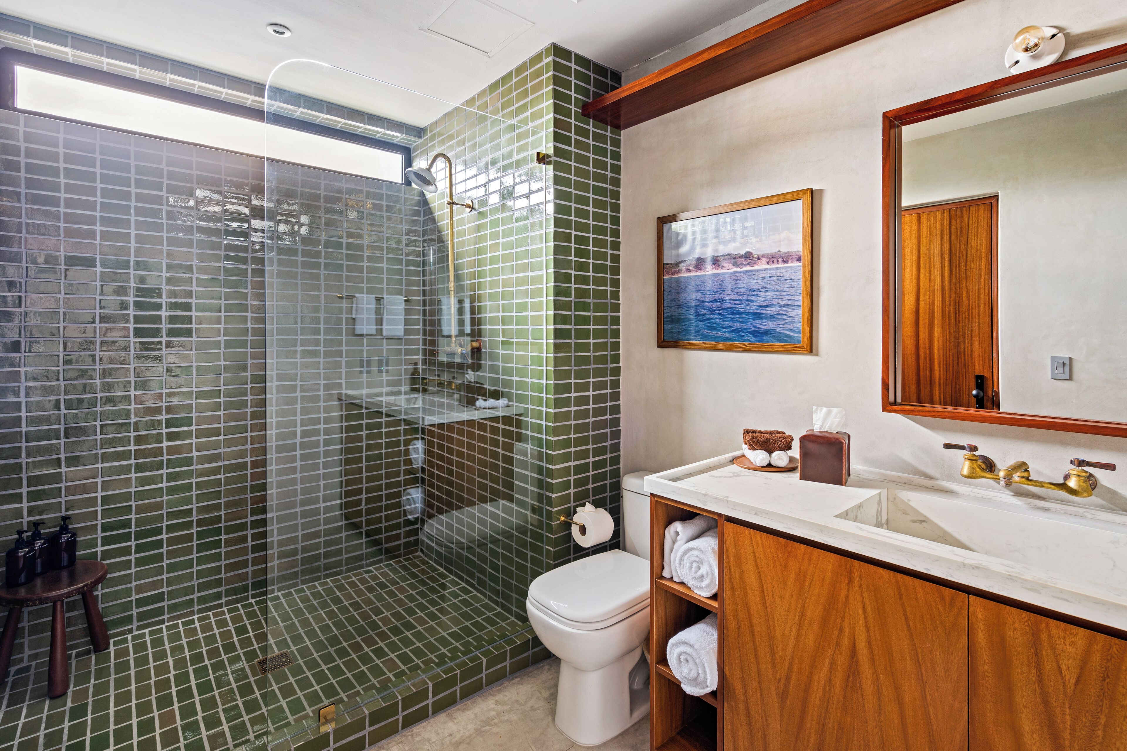 Een badkamer met een douche met glazen wand, een toilet, een wastafel met houten kastje, en een ingelijst schilderij aan de muur.