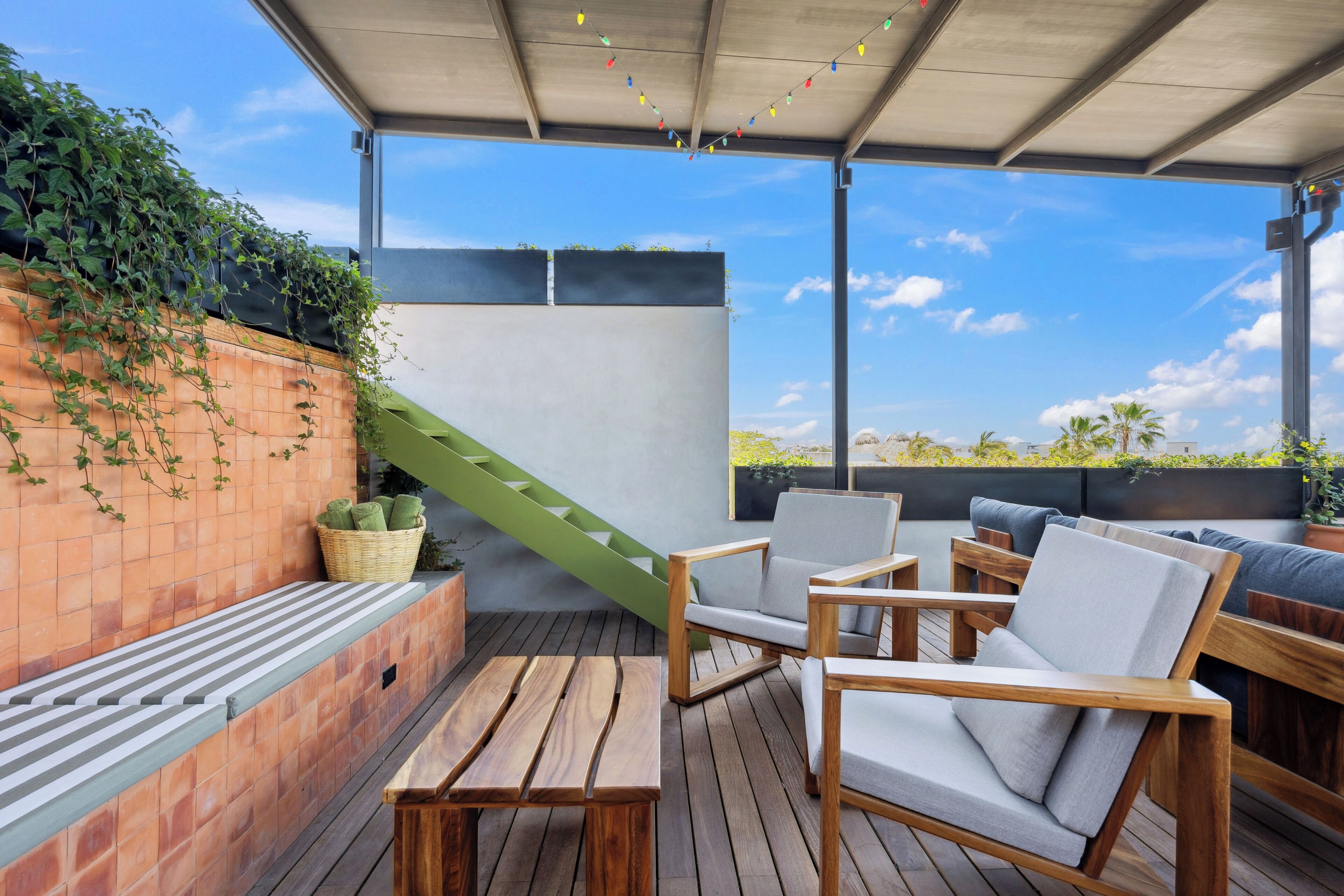 Un espacio de estar al aire libre cubierto, con mobiliario de madera, una escalera verde y vistas a palmeras.