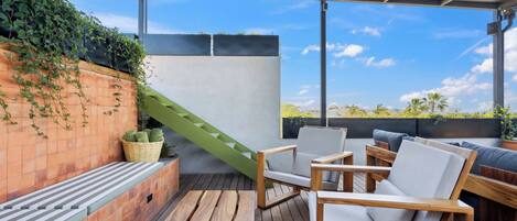 Un espacio de estar al aire libre cubierto, con mobiliario de madera, una escalera verde y vistas a palmeras.