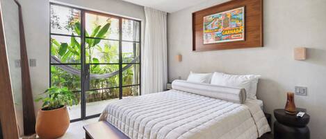 A bedroom with a large window, a bed, a bench, a potted plant, and a framed picture on the wall.