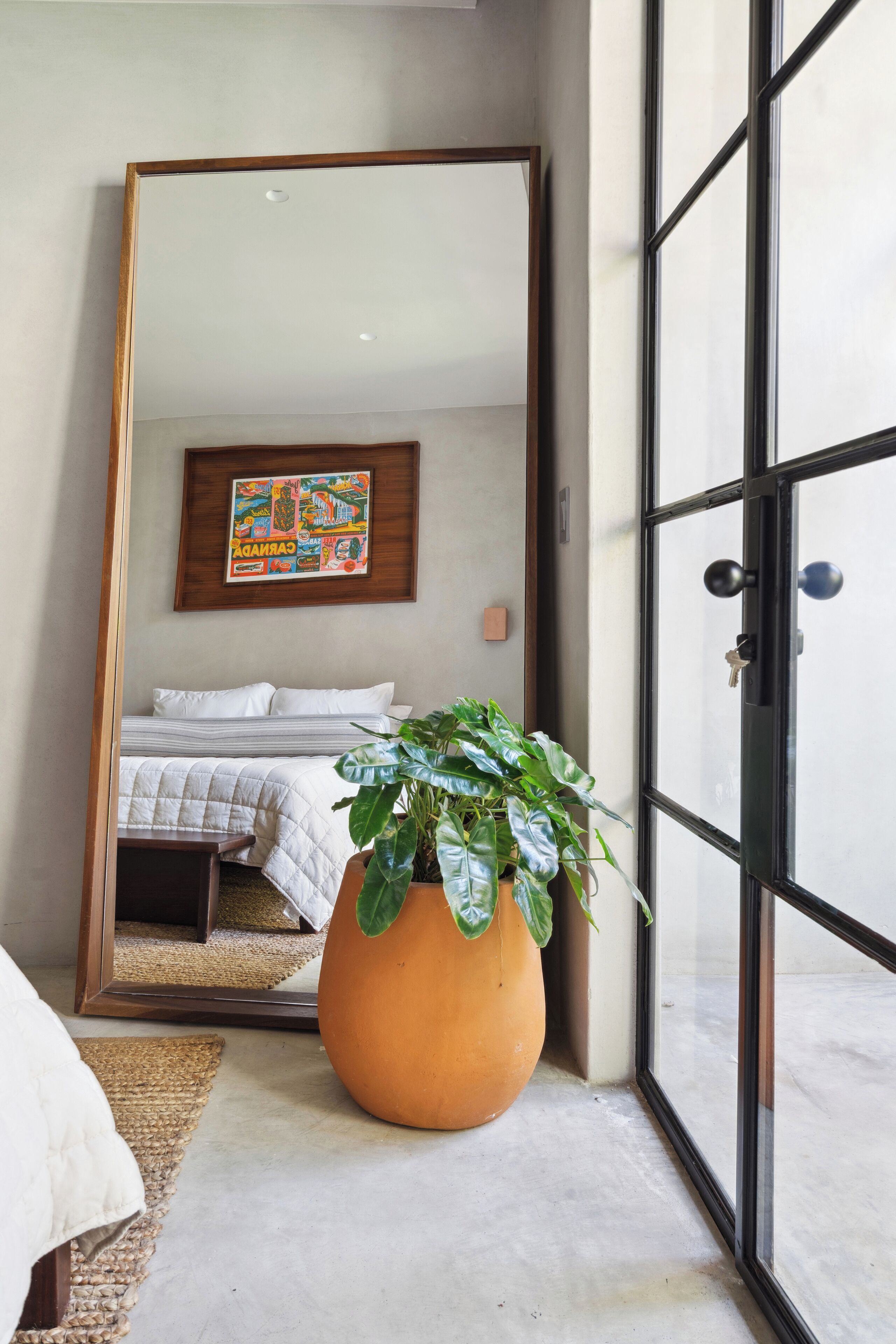 Un dormitorio con un espejo grande que refleja una cama, una planta en maceta y una obra de arte colgada en la pared.