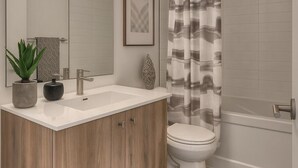 Een moderne badkamer met een houten wastafelmeubel, een witte wasbak, een lijstfoto, een potplant en een douche met een decoratieve gordijn.