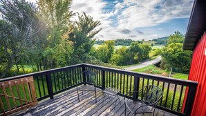 Una terraza de madera con sillas y una mesa de metal, con vistas a un paisaje verde con árboles y una carretera.