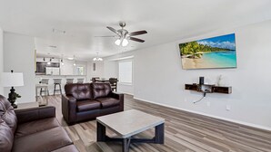 A modern living room with a brown leather sofa, a coffee table, and a ceiling fan. There is a kitchen in the background with white cabinets and a dining area with a table and chairs.