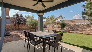 An outdoor patio with a table and chairs, a ceiling fan, and a view of a landscaped garden and stone wall.
