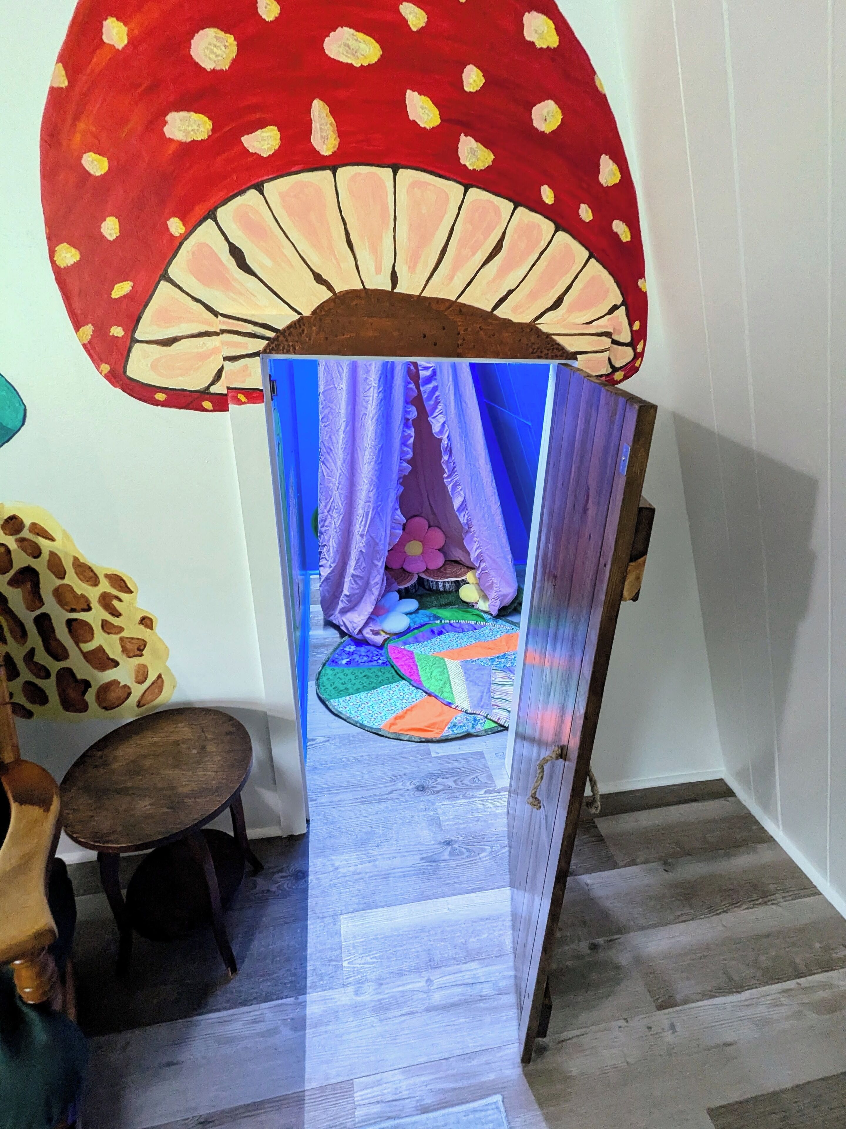 A room with a large mushroom mural on the wall, a bed with a colorful quilt, and a small wooden stool.