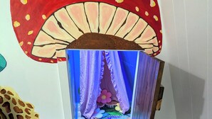 Una habitación con un gran mural de hongos en la pared, una cama con un edredón colorido y un pequeño taburete de madera.