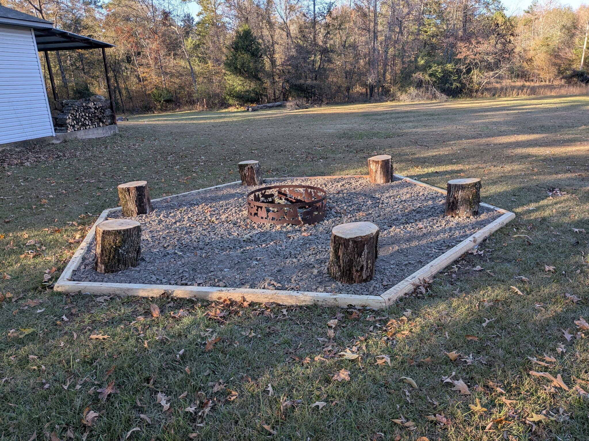 A circular fire pit with a metal grate, surrounded by a gravel bed and bordered by wooden stumps.
