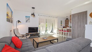 Una sala moderna con un sofá gris, una mesa de centro y un televisor. Se observa un comedor con sillas blancas y una cocina al fondo.