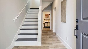 Un escalier aux murs blancs et aux marches sombres, menant à une pièce où se trouvent un lit et un tableau accroché au mur.