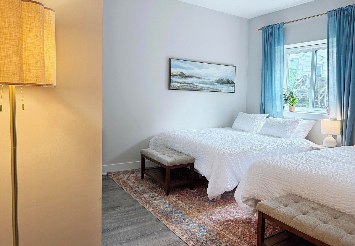 A bedroom with two beds, a bedside table, a lamp, a painting on the wall, and a window with curtains.