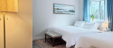 A bedroom with two beds, a bedside table, a lamp, a painting on the wall, and a window with curtains.