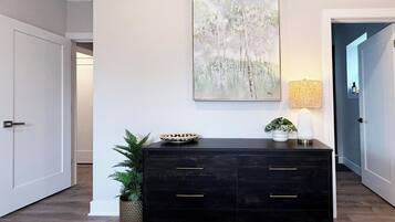 A hallway with a dark wooden dresser, a wall-mounted abstract painting, a potted plant, and a lamp.