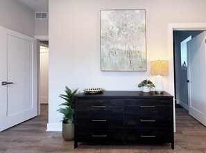 A hallway with a dark wooden dresser, a wall-mounted abstract painting, a potted plant, and a lamp.
