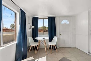 Une pièce avec une porte blanche, une fenêtre ornée de rideaux bleus, une table ronde avec deux chaises et un sol carrelé.