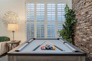 A pool table with balls, a lamp, a window with blinds, and a plant.
