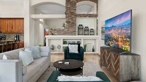 A living room with a sofa, coffee table, and a TV. There is a kitchen area with cabinets and a dining table. A fireplace is visible in the background. - Lakeside home with private patio, Grill, Golf view (Glendale)