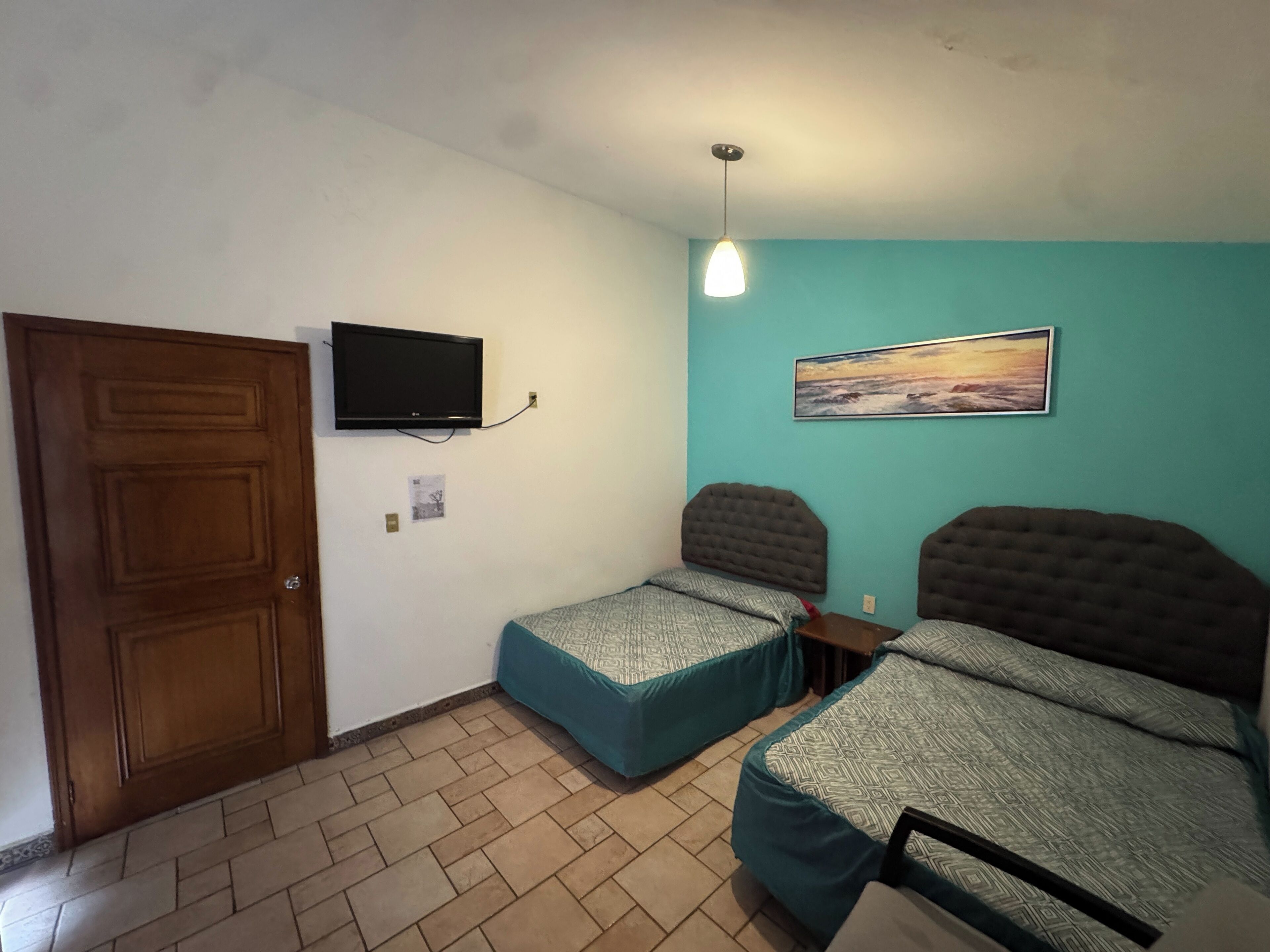 A room with two beds, a television mounted on the wall, a painting, and a wooden door.