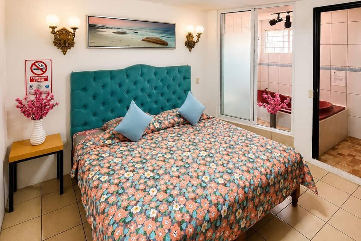 A bedroom with a teal headboard, floral bedspread, a painting, a vase with flowers, and a view of a bathroom through a glass door.
