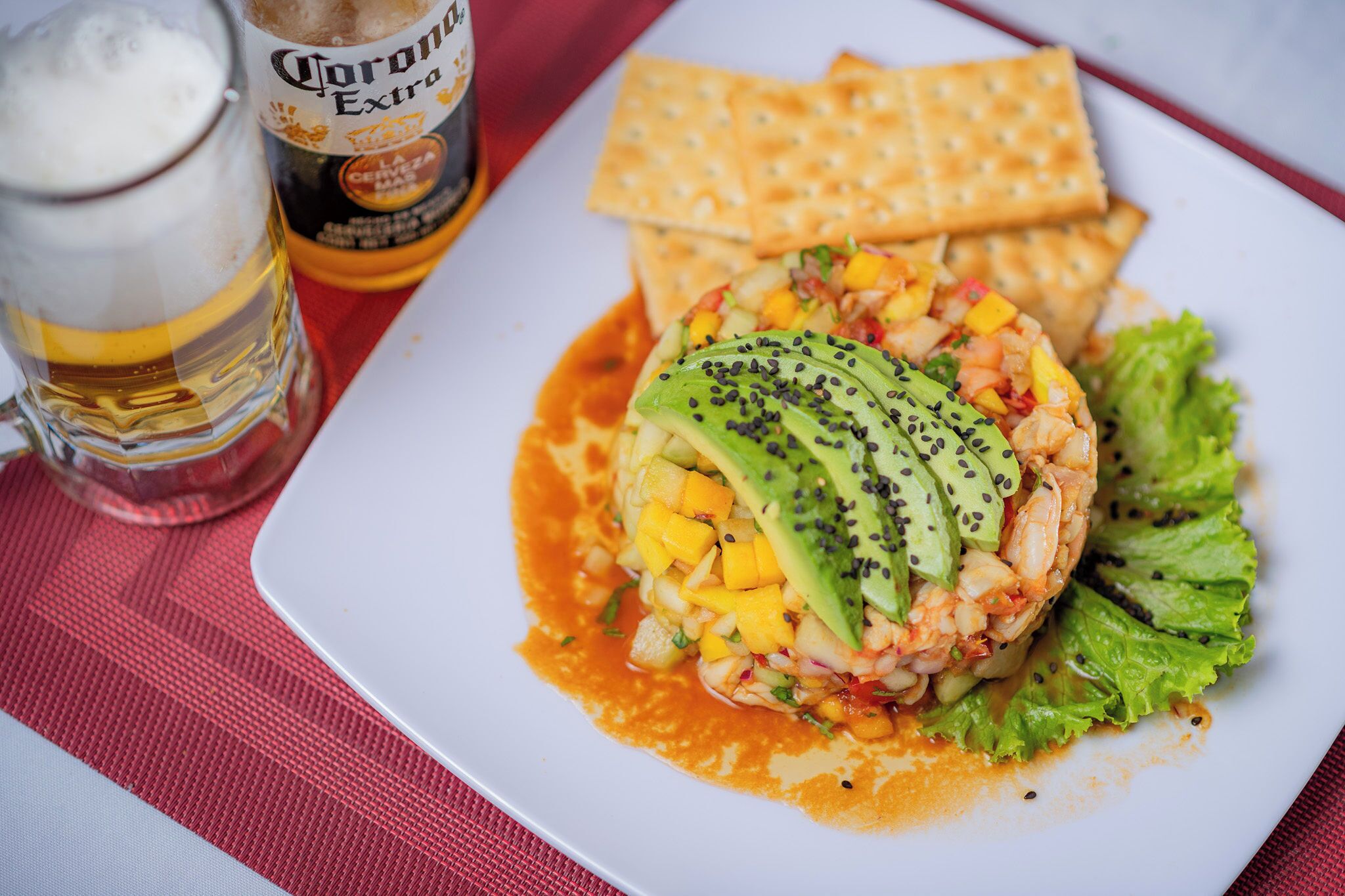 Un plato con ensalada de camarones, aguacate y mango, una jarra de cerveza y galletas sobre un mantel rojo.