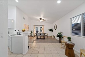 A modern kitchen with white cabinetry, a dining area with a table and chairs, a ceiling fan, and a window with blinds.
