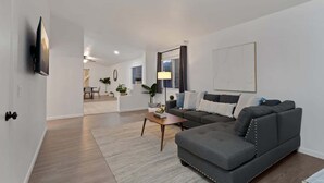 Ein modernes Wohnzimmer mit einem grauen Ecksofa, einem Couchtisch, einem an der Wand befestigten Flachbildfernseher und einem Essbereich im Hintergrund.