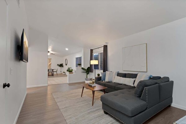 A modern living room with a grey sectional sofa, a coffee table, a flat-screen TV mounted on the wall, and a dining area in the background.