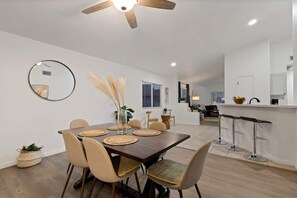 A dining area with a wooden table, chairs, a ceiling fan, and a kitchen in the background.