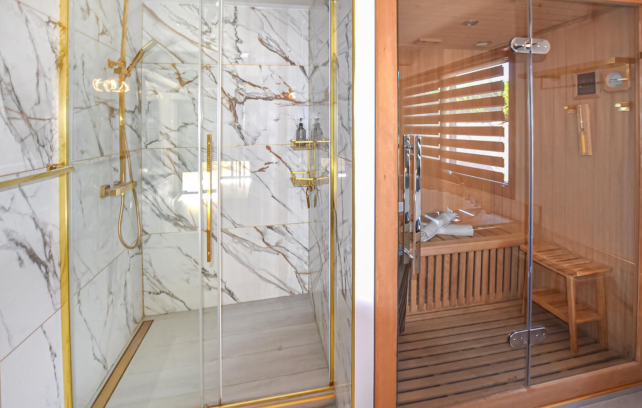 A glass-enclosed shower with gold fixtures and marble walls, and a wooden sauna with benches and a glass door.