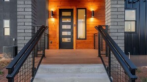 A modern building with a wooden front porch, a black door, and two wall-mounted lights. - The Bluebird Flagstaff, Jacuzzi, Sleeps 26 (Flagstaff)