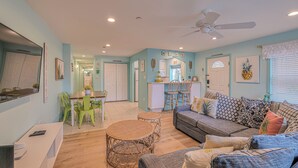 A spacious living room with a grey sofa, a dining table with chairs, a ceiling fan, and a kitchen area in the background. - Pineapple Pad:ACROSS from beach + boardwalk + parking w/ Full Linen Service! (Ocean City)