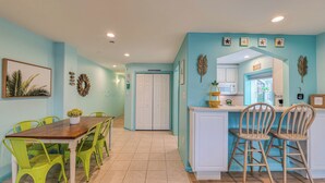 A dining area with a wooden table and green chairs, a kitchen with white cabinets and a window, and a hallway with a white door. - Pineapple Pad:ACROSS from beach + boardwalk + parking w/ Full Linen Service! (Ocean City)