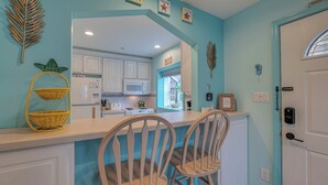 A kitchen with white cabinets, a microwave, and a window with a view of the outside. There are two chairs and a yellow wicker shelf with baskets. - Pineapple Pad:ACROSS from beach + boardwalk + parking w/ Full Linen Service! (Ocean City)