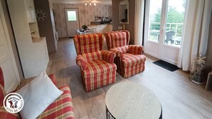 Uma sala de estar com sofá, poltronas e mesa de centro. Ao fundo, uma área de jantar com mesa e cadeiras.