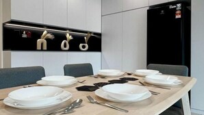 Un espace repas moderne avec une table en bois dressée pour six personnes, un réfrigérateur noir et une étagère murale agrémentée d’objets décoratifs.