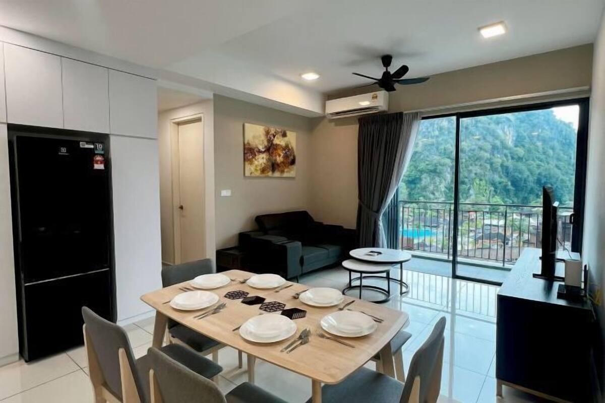Un coin repas avec une table dressée pour quatre personnes, un réfrigérateur noir, un canapé gris, une petite table ronde et un balcon avec vue sur la verdure.