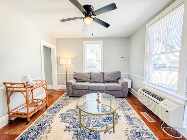 A living room with a grey sofa, a glass coffee table, a wicker side table, a ceiling fan, and a large window with blinds.
