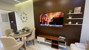 Un salon moderne avec un téléviseur à écran plat fixé à un mur en bois, une table à manger avec des chaises, un canapé et une petite étagère ornée d’objets décoratifs.