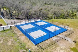 Een groot tennisveld buiten met een blauwe oppervlakte en witte lijnen, omheind door een metalen hek.