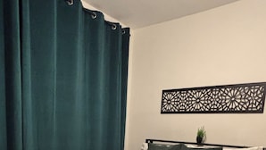 Ein ordentlich bezogenes Bett mit einer weiß-grünen Tagesdecke, einem Kissen und einem gefalteten weißen Handtuch. Darüber an der Wand hängt eine schwarze Dekoration.